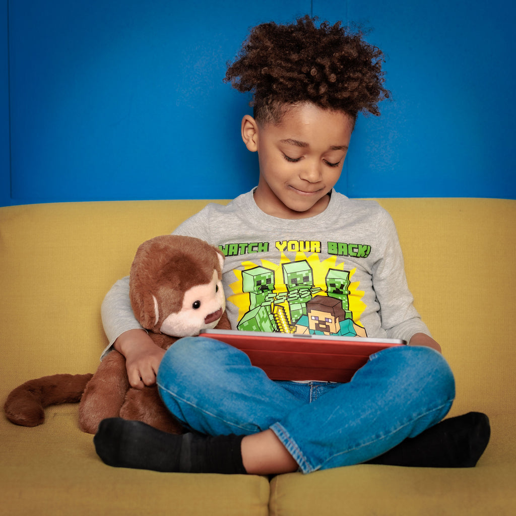 Boy on a couch holding Motivational Monkey and a tablet, with the toy encouraging play and learning.