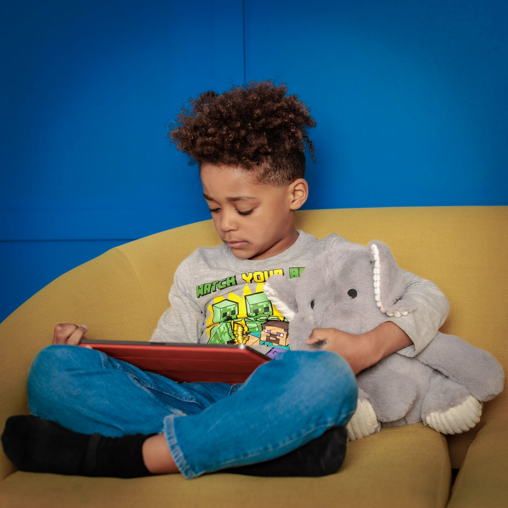 Boy on a couch holding Empowerment Elephant, absorbed in learning through a tablet app.