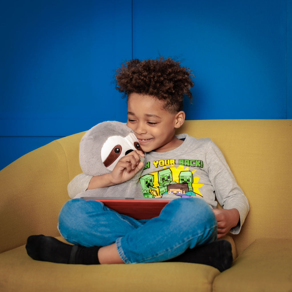 Smiling child gently holding Tranquil Sloth while reading on the couch, feeling soothed and comforted.