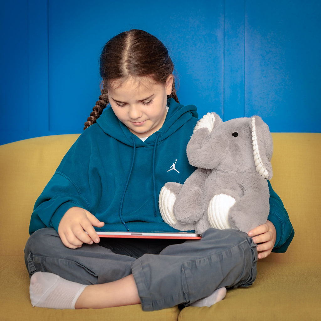 Girl with Empowerment Elephant on a couch, using a tablet, with the toy supporting her educational journey.