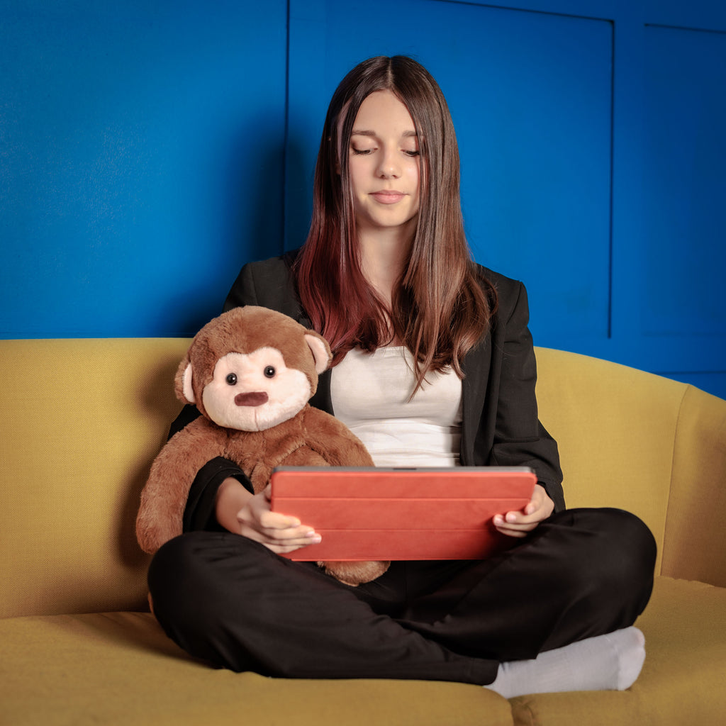 Girl and Motivational Monkey on a couch, the toy sitting upright as she uses a learning app on a tablet.